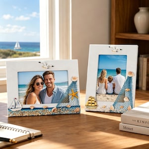 May include: Two white rectangular picture frames decorated with nautical elements including a blue fishing net, seashells, a starfish, a sailboat, and a seagull. One frame is horizontal and the other is vertical, both displaying beach photos.