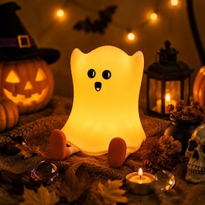 May include: A glowing, warm yellow ghost-shaped lamp with a smooth, rounded body, two black oval eyes, and an open mouth. The lamp features two small, orange, foot-like protrusions extending from the base, creating a seated appearance.