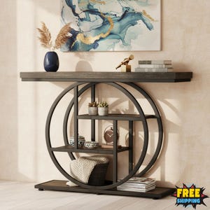 May include: A modern console table with a dark wood top and a black metal circular frame. The table features multiple shelves for storage and display, with decorative items such as plants, books, and a clock. A painting with blue and gold accents hangs above.