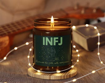 May include: A lit brown glass candle jar with the letters "INFJ" in green on the label. The label also features a drawing of a person and the words "Illuminate the path with compassion". The candle sits on a wooden coaster, with string lights around it.