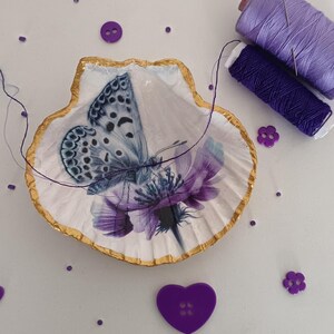 Può includere: Un piatto a conchiglia con un disegno di farfalla e fiori, bordi dorati e accenti viola. L'immagine include rocchetti di filo viola, bottoni e abbellimenti di bottoni a forma di cuore. Tema artigianale.
