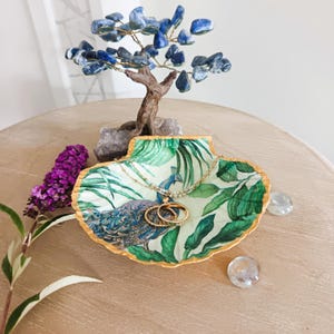 Peut inclure: Un vide-poche en forme de coquille Saint-Jacques orné d'un motif de paon et de feuilles tropicales à l'intérieur, avec un bord festonné peint en doré. Le plat contient deux bagues en métal doré et un collier délicat en perles.