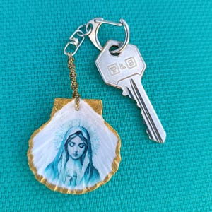 Peut inclure: Un porte-clés artisanal avec un pendentif en coquillage peint représentant une figure religieuse en bleu et blanc. La coquille est bordée de peinture dorée et attachée à un porte-clés argenté avec une clé.