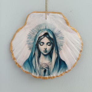 Può includere: Un ornamento decorativo da appendere realizzato con una conchiglia di capasanta bianca, raffigurante un'illustrazione di una figura con occhi chiusi e mani giunte in preghiera, con un velo blu, bordo dorato e un cordino per appenderlo.