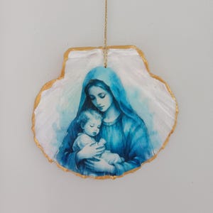 Puede incluir: Un adorno colgante decorativo hecho de una concha de vieira blanca con una ilustración en tonos azules de una figura materna sosteniendo a un niño. La concha tiene un borde pintado en dorado y cuelga de un cordón fino de color oro.