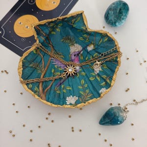 Peut inclure: Un collier doré avec un pendentif en forme d'étoile est posé sur un plat décoratif turquoise avec un motif de colibri et de fleurs. Un pendentif en pierre bleue et une pierre bleue sont également présents.