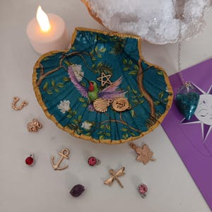 Può includere: Un vassoio portagioie a forma di conchiglia turchese con bordo dorato, con un disegno di colibrì. Vari ciondoli dorati, tra cui una stella, una conchiglia e una foglia, sono sparsi intorno al vassoio. Sono presenti anche una candela e un pendolo.