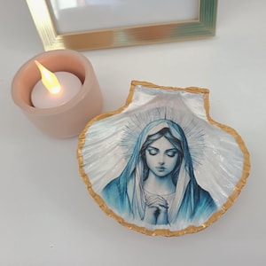Può includere: Conchiglia decorativa con bordo dorato, raffigurante l'immagine di una donna con velo blu e aureola. Accanto, un piccolo portacandele rosa con candela accesa.