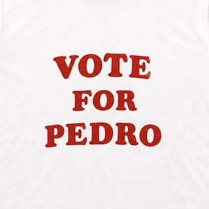 Op de afbeelding: Een wit ringer-t-shirt met zwarte boorden aan de kraag en mouwen. Op de voorkant van het shirt staat de tekst VOTE FOR PEDRO gedrukt in dikke, rode hoofdletters, gecentreerd op het borstgedeelte van het kledingstuk.