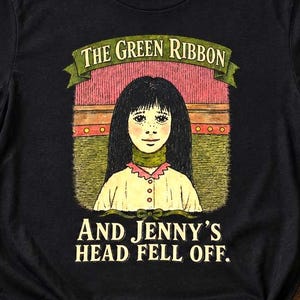 Puede incluir: Una camiseta negra de manga corta con una ilustración gráfica de una persona con cabello largo y oscuro que lleva una cinta verde alrededor del cuello. El texto THE GREEN RIBBON AND JENNY'S HEAD FELL OFF aparece en una fuente estilizada.