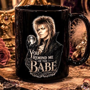 May include: A black ceramic mug featuring a graphic of a person with long hair holding a crystal ball, with the text "YOU REMIND ME OF THE BABE" printed in a stylized white font below the image. The mug has a standard handle and a glossy finish.
