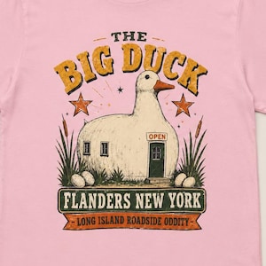 May include: A light pink t-shirt featuring a graphic of The Big Duck building in Flanders, New York. The design includes the text THE BIG DUCK, FLANDERS NEW YORK, and LONG ISLAND ROADSIDE ODDITY, with illustrations of stars, reeds, and eggs.