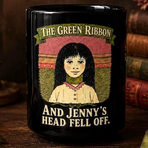 May include: A black ceramic mug featuring a central illustration of a person with long dark hair wearing a green ribbon around their neck. The text above reads THE GREEN RIBBON and the text below reads AND JENNY'S HEAD FELL OFF.