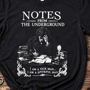 May include: A black short-sleeved t-shirt featuring a white graphic print of a person writing at a desk by candlelight. The text reads NOTES FROM THE UNDERGROUND above the image and I AM A SICK MAN... I AM A SPITEFUL MAN below the image.