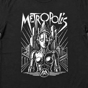 Puede incluir: Una camiseta negra de manga corta con un estampado gráfico blanco de una figura robótica frente a un horizonte urbano estilizado. El texto "METROPOLIS" está impreso en una fuente angular sobre el robot, con un logotipo circular.