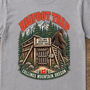Puede incluir: Una camiseta gris de manga corta con un gráfico de una estructura de madera etiquetada como Collings Mountain Bigfoot Trap, una figura de Bigfoot, pinos y montañas. El texto dice Bigfoot Trap y Collings Mountain, Oregon, con una pequeña bandera.