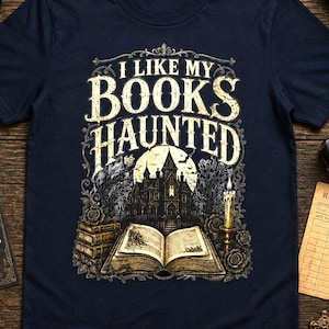 May include: A navy blue short-sleeve t-shirt featuring a graphic print of a haunted mansion, an open book, and a lit candle. The text "I LIKE MY BOOKS HAUNTED" is printed in a decorative, weathered white font above the central illustration.