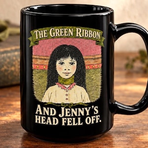 Puede incluir: Una taza de cerámica negra con una ilustración gráfica de una persona con cabello largo y oscuro que lleva una cinta verde alrededor del cuello. El texto dice THE GREEN RIBBON arriba y AND JENNY'S HEAD FELL OFF abajo en letras blancas.