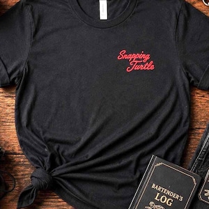 Peut inclure: T-shirt noir avec "Snapping Turtle" en script rouge, associé à un jean noir, des bottes et des accessoires. Comprend un dessous de verre, une bougie, un carnet de barman et un porte-clés "Support Local Dive Bars".
