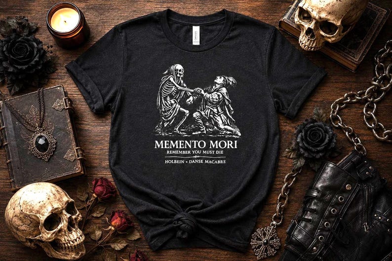 May include: A black short-sleeved t-shirt featuring a white graphic print of a skeleton and a person kneeling. The text reads MEMENTO MORI, REMEMBER YOU MUST DIE, and HOLBEIN DANSE MACABRE. The shirt has a crew neckline and a standard fit.