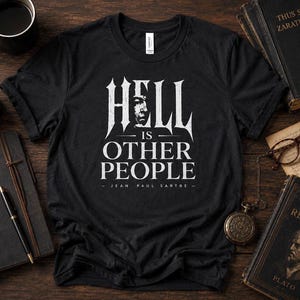 Puede incluir: Camiseta negra con la frase "HELL IS OTHER PEOPLE" y un retrato, atribuida a Jean-Paul Sartre. La camiseta se muestra sobre una superficie de madera con libros, una vela, una taza de café y un cuaderno.