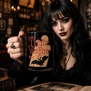 Pode incluir: Uma caneca de cer&acirc;mica preta com o texto vermelho Theatre des Vampires, uma silhueta de lua bege e um gr&aacute;fico de um morcego preto. A caneca tem uma al&ccedil;a resistente e um acabamento liso, exibindo o texto La mort en chantant sob a ilustra&ccedil;&atilde;o.