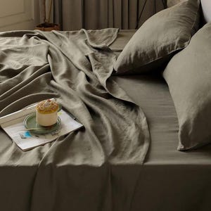 May include: A bed with olive green linen bedding, including a flat sheet, fitted sheet, and two pillows. A small dessert sits on a green plate atop a magazine. A paper lantern hangs above the bed.