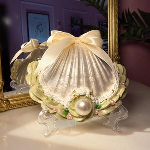 Puede incluir: Adorno decorativo en forma de concha con una perla grande y un lazo color crema. La concha plateada se asienta sobre una base verde claro con detalles de perlas rosas y pequeñas rosas. El adorno está sostenido por soportes de plástico transparente.