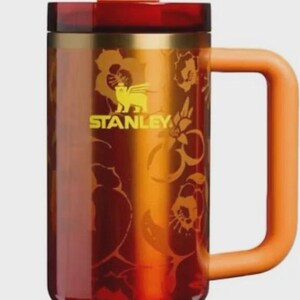 K&ouml;nnte beinhalten: Ein Stanley-Reisebecher mit einem goldenen Ananas-Aufsatz und einem orangefarbenen Griff. Der Becher hat ein florales Design und das Stanley-Logo. Der Farbverlauf geht von Rot zu Orange &uuml;ber.