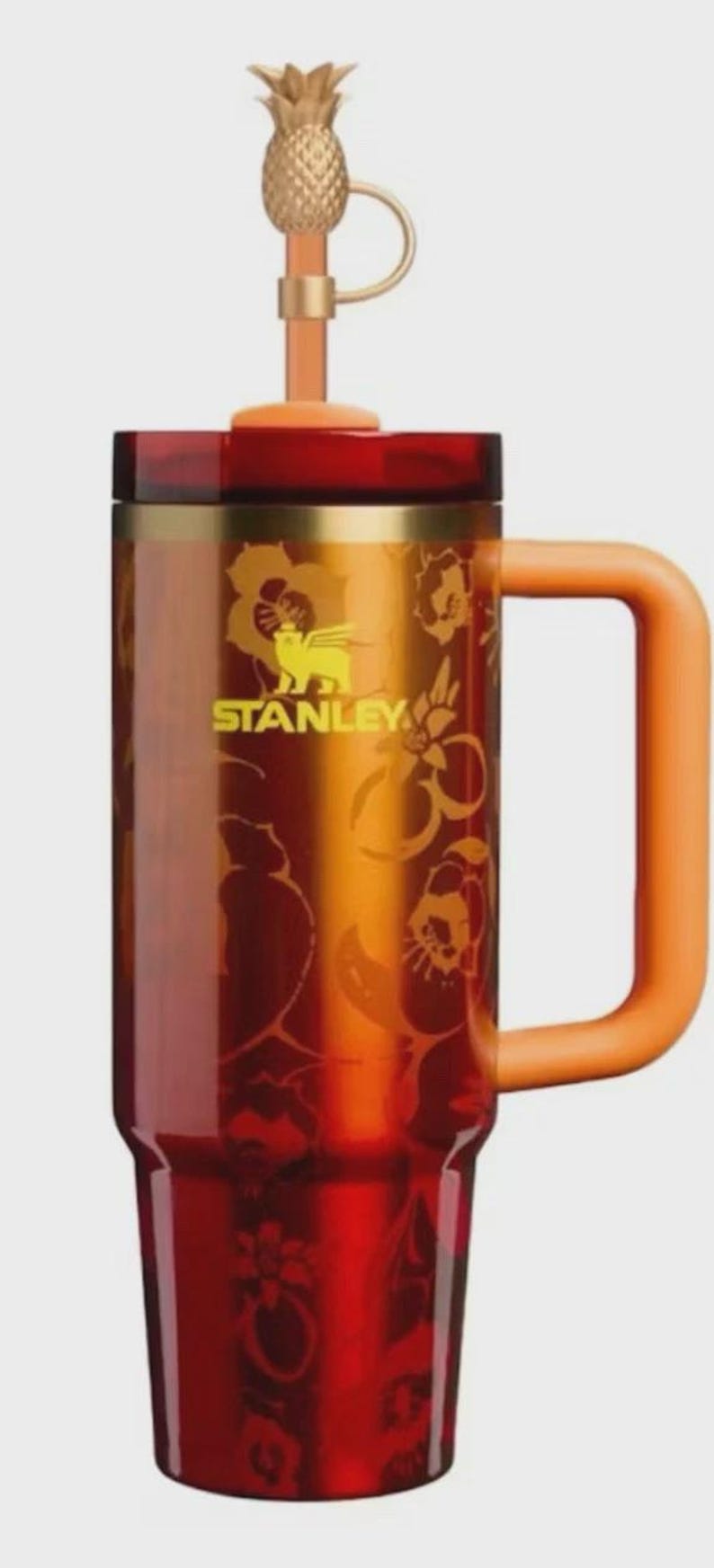 K&ouml;nnte beinhalten: Ein Stanley-Reisebecher mit einem goldenen Ananas-Aufsatz und einem orangefarbenen Griff. Der Becher hat ein florales Design und das Stanley-Logo. Der Farbverlauf geht von Rot zu Orange &uuml;ber.