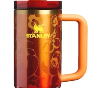 K&ouml;nnte beinhalten: Ein Stanley-Reisebecher mit einem goldenen Ananas-Strohhalmaufsatz und einem orangefarbenen Griff. Der Becher hat eine rot-goldene Farbverlaufsoptik mit floralen Details und Logo. Das Stanley-Logo ist sichtbar.