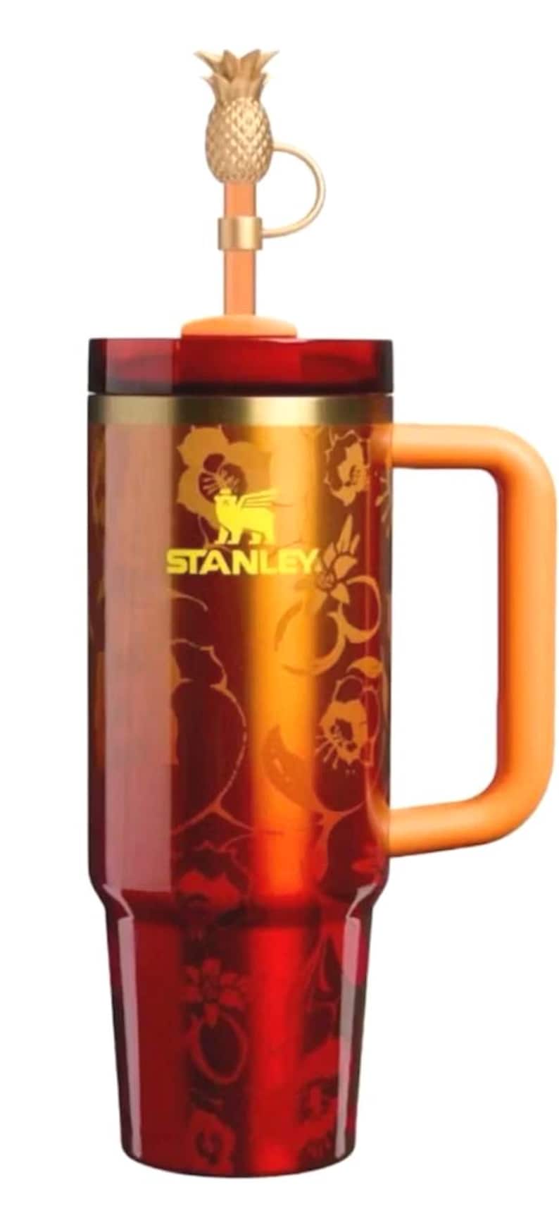K&ouml;nnte beinhalten: Ein Stanley-Reisebecher mit einem goldenen Ananas-Strohhalmaufsatz und einem orangefarbenen Griff. Der Becher hat eine rot-goldene Farbverlaufsoptik mit floralen Details und Logo. Das Stanley-Logo ist sichtbar.