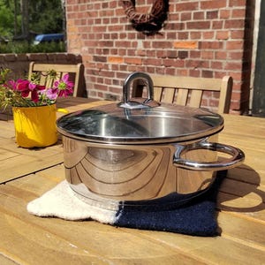 May include: A polished stainless steel cooking pot with a matching lid, featuring two rounded side handles and a central arched handle on the lid. The pot sits on a rectangular, two-toned white and navy blue felt trivet on a wooden surface.