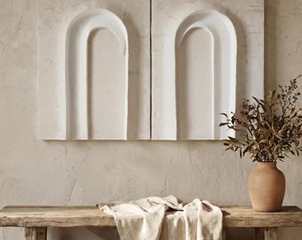 Puede incluir: Dos paneles de pared rectangulares de color blanco roto con un diseño de arco empotrado, un banco de madera rústica con una tela clara drapeada y un jarrón de color terracota con ramas secas, todo dispuesto en una exhibición coherente.