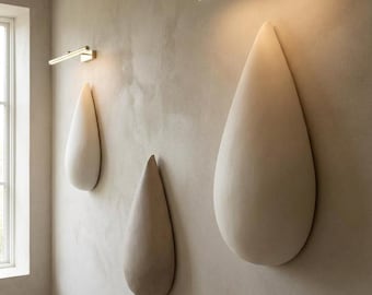 Puede incluir: Tres esculturas de pared en forma de gota de color blanco roto montadas sobre una pared beige texturizada. Cada escultura está iluminada desde arriba por una lámpara de pared horizontal y delgada de latón cepillado con luz cálida.