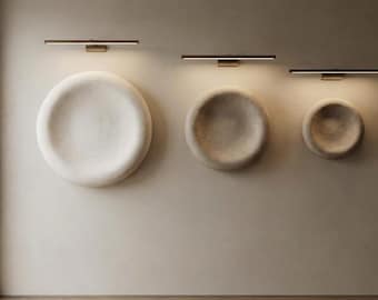 Puede incluir: Tres esculturas de pared circulares en forma de cuenco de diferentes tamaños, dispuestas horizontalmente. Cada pieza es de color blanco roto con una superficie texturizada e iluminada desde arriba por una lámpara de metal oscuro, fina y horizontal.