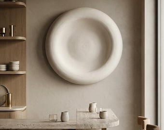 Puede incluir: Una gran escultura de pared circular de color blanco roto con un centro cóncavo montada en una pared de tonos neutros. La habitación cuenta con una encimera de piedra, estanterías de madera con varios recipientes y sillas tapizadas.