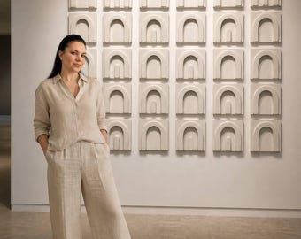 Puede incluir: Una mujer posa frente a una instalación de arte mural. El arte consiste en múltiples paneles cuadrados, cada uno con un diseño arqueado repetido en un color neutro. La mujer viste una camisa de lino beige y pantalones anchos a juego.