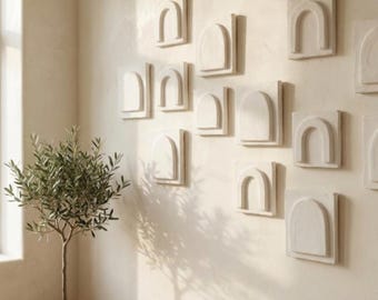 Puede incluir: Una exhibición mural de piezas de arte de pared arqueadas de color blanquecino. El arte está dispuesto en un patrón de cuadrícula sobre una pared de color beige claro. Un pequeño olivo en maceta y un banco de madera con una manta beige están en primer plano.