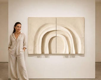 Puede incluir: Una escultura abstracta en relieve beige con formas curvas y en capas está montada en una pared blanca. La obra de arte está dividida en dos paneles. Una persona con un atuendo de lino beige está de pie junto a la obra de arte.