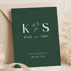 Può includere: Un invito di nozze verde scuro con il testo bianco K e S separati da un disegno al tratto di un rametto, con i nomi Kevin e Sara, la frase A love story begins anew e la data 21st July 2026 stampati in basso.