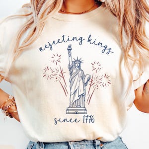 May include: An ivory t-shirt featuring a dark blue graphic of the Statue of Liberty surrounded by red fireworks. The text above the statue reads "rejecting kings" and the text below reads "since 1776" in a dark blue cursive script font.