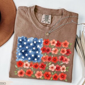May include: Espresso-colored t-shirt featuring a floral American flag design. The flag design includes a blue watercolor square with white stars and rows of red and pink flowers. The shirt has a crew neck and short sleeves.