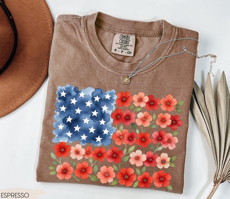 May include: Espresso-colored t-shirt featuring a floral American flag design. The flag design includes a blue watercolor square with white stars and rows of red and pink flowers. The shirt has a crew neck and short sleeves.