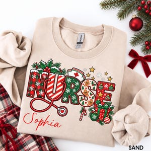 May include: A sand-colored crewneck sweatshirt featuring a graphic print of the word NURSE in festive patterns, including red plaid, candy cane stripes, and gingerbread men. A red stethoscope, holly, and a gift box are integrated into the design.