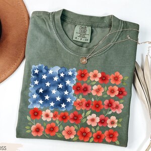 May include: A moss green t-shirt featuring a floral American flag design. The flag design includes a blue watercolor square with white stars and rows of red and pink flowers. The shirt has a crew neck and short sleeves.
