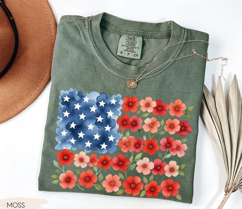 May include: A moss green t-shirt featuring a floral American flag design. The flag design includes a blue watercolor square with white stars and rows of red and pink flowers. The shirt has a crew neck and short sleeves.