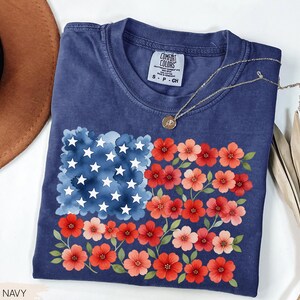 May include: A navy blue t-shirt featuring a floral American flag design. The flag's stars are white, the stripes are red and pink flowers, and the background is a watercolor blue. The shirt has a Comfort Colors tag.