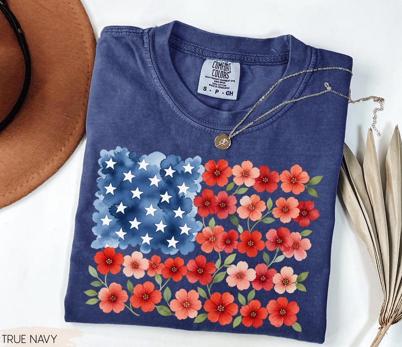 May include: A navy blue t-shirt featuring a floral American flag design. The flag's stars are white, the stripes are red and pink flowers, and the background is a watercolor blue. The shirt has a Comfort Colors tag.