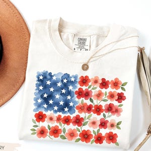 May include: Ivory-colored t-shirt featuring a watercolor design of the American flag, with blue stars and red and pink flowers. The shirt has a crew neck and short sleeves. A gold necklace with a pendant is draped across the shirt.